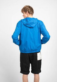 Blå vindjakke med hætte, elastiske manchetter og talje; parret med sorte shorts med en synlig label på baglommen.