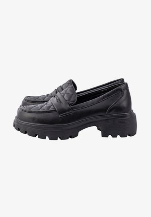 Personne portant un pantalon rose taille haute à jambes larges et des mocassins noirs épais, posant avec une jambe levée et la main sur la hanche sur un fond uni.