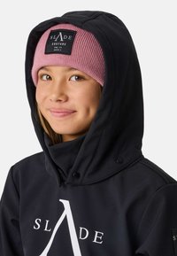 Giacca nera con cappuccio e logo bianco, caratterizzata da un collo alto e bottoni a scatto. Sopra è posizionato un berretto rosa a costine con un'etichetta nera.