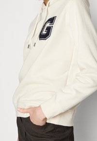 Sudadera con capucha color crema con un logo "G" en azul marino, capucha con cordón y puños acanalados. Hecha de tela suave con un ajuste cómodo.