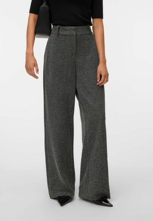 Femme portant un pantalon large taille haute gris à motif chevrons, des chaussures noires pointues, un top noir à manches courtes, et un sac à main noir.