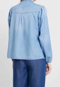 Vaaleansininen farkkupaita, jossa on kerätty pääntie, pitkät hihat joustavilla hihoilla ja rento leikkaus, takana on laskostusyksityiskohta.