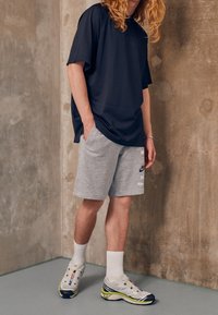 Svart mesh t-shirt med kort ärm, grå shorts med tryck, vita strumpor och flerfärgade sportskor, stående på ett slätt grått golv.