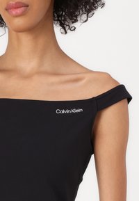 Juodas ofšoulder paaukštintas viršus, pagamintas iš lygaus audinio. Turintis baltą "Calvin Klein" logotipą ant krūtinės ir trumpas rankoves su suktomis pakraštėmis.