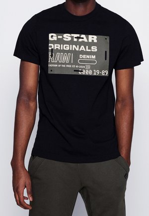 Mężczyzna noszący czarną koszulkę G-Star Originals z białym i szarym graficznym tekstem, w połączeniu z ciemnooliwkowymi spodniami, z rękami w kieszeniach.