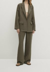 Olijfgroene blazer en bijpassende wijde broek van textuurstof. De blazer heeft twee voorzakken en een relaxte pasvorm.