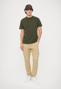 T-shirt verde de mangas curtas com um pequeno logotipo, combinado com calças cargo bege claro com vários bolsos, usadas com sapatilhas brancas.