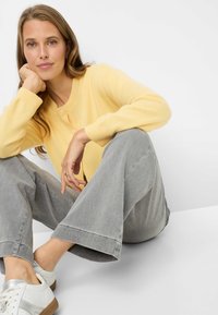 Cardigan giallo con bottoni, realizzato in tessuto morbido; abbinato a jeans grigio chiaro a gamba larga e sneaker bianche con dettagli argentati.