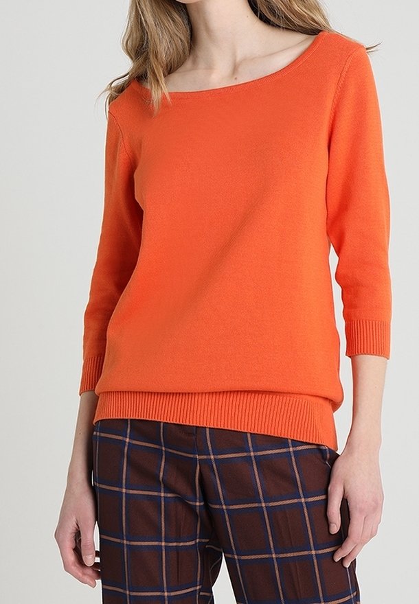Femme portant un pull orange vif à col bateau avec des manches trois-quarts et un pantalon à carreaux marron avec des lignes bleues et orange.