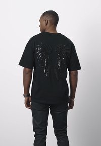 Musta lyhythihainen t-paita, jossa on paljeteilla koristeltu siipikuvio takana. Mallilla on mustat housut, joissa on rento istuvuus.