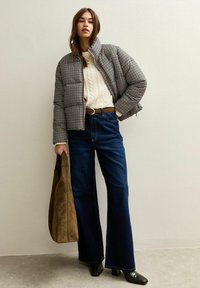 Chaqueta acolchada con estampado de pata de gallo, suéter de punto trenzado color crema, jeans oscuros de pierna ancha, botas negras de tacón y un bolso de hombro marrón.