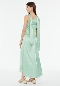 Abito lungo in satin verde menta con corpetto aderente, schiena scoperta e due lunghi nastri che cadono. Presenta una gonna fluida e un hardware minimale.