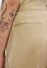 Τατουαρισμένο χέρι με διάφορα μελανά πρόσωπα και σχέδια δίπλα σε μπεζ παντελόνι με οριζόντια πίσω τσέπη.
