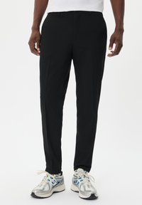 Pantalones de vestir negros con un corte ajustado, frente plano y una textura de tela suave, combinados con zapatillas deportivas grises y azules.