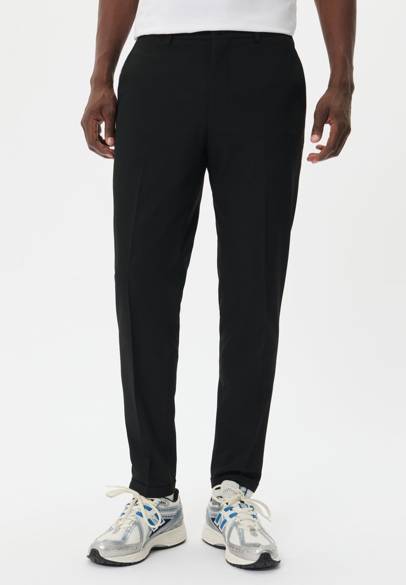 Pantalones de vestir negros con un corte ajustado, frente plano y una textura de tela suave, combinados con zapatillas deportivas grises y azules.