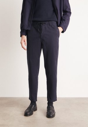 Persona che indossa pantaloni blu navy con coulisse, calzini neri, mocassini in pelle neri e una giacca blu navy abbinata con la mano in tasca.