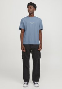Camiseta de algodón azul claro con mangas cortas y cuello redondo. Presenta un texto de logo blanco en el frontal. Combinada con pantalones cargo negros y zapatillas.