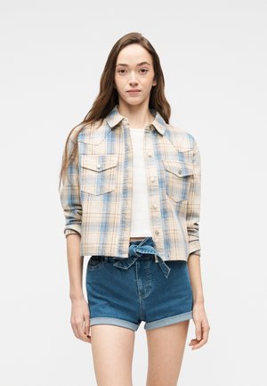 Νεαρή γυναίκα φοράει ένα πουκάμισο με μπεζ και μπλε καρό κουμπωτό πάνω από λευκό τοπ, συνδυασμένο με τζιν σορτς με ρολά και ζώνη στη μέση.