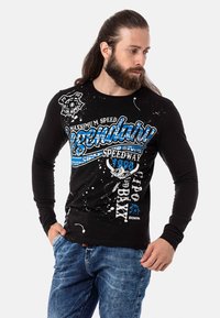 Schwarzes langärmliges Shirt mit blau-weißer Grafik, Splattermustern und Rundhalsausschnitt. Getragen mit blauen Jeans.