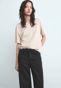 Beige T-shirt met korte mouwen, relaxte pasvorm, gedragen met hoge zwarte jeans. De stof lijkt zacht met een gladde textuur.