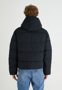 Chaqueta acolchada negra con capucha, con costuras horizontales y un corte corto, combinada con vaqueros de mezclilla azules.