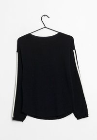 Maglione nero a maniche lunghe con strisce bianche sulle maniche appeso a un gancio nero contro un muro bianco.