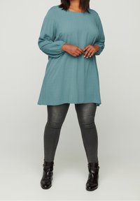 Haut à manches longues couleur sarcelle avec une finition texturée, présentant une coupe décontractée et des épaules tombantes, associé à un jean slim gris foncé et des bottines noires.