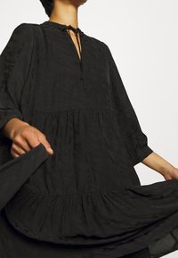 Vestido negro en capas con escote redondo y cierre con lazo. Hecho de un material ligero, con mangas largas y una textura sutil.