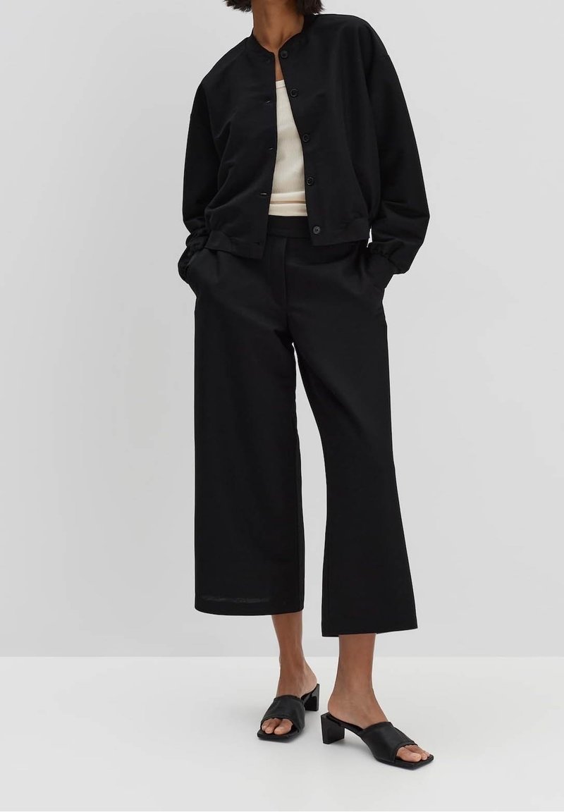 Femme portant une veste noire boutonnée, un haut blanc, un pantalon noir large coupé court, et des sandales noires à talons ouverts avec talons carrés.