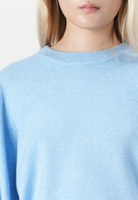 Detailní záběr osoby v světle modrém pleteném svetru s kulatým výstřihem a dlouhými blond vlasy na bílém pozadí.
