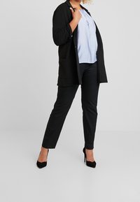 Czarny długi kardigan z dwoma przednimi kieszeniami, noszony na jasnoniebieskiej bluzce i czarnych obcisłych spodniach, uzupełniony czarnymi szpilkami.