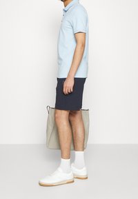 Polo azzurro chiaro, pantaloni corti blu navy, sneakers bianche con suole in gomma e borsa grigia. Presenta maniche corte e un dettaglio logo discreto.