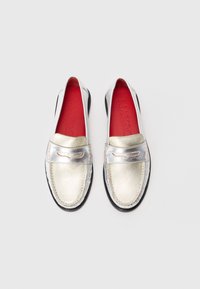 Metallic silver slip-on shoes with a smooth texture, featuring a red insole and black rubber sole. Accented with a decorative strap.