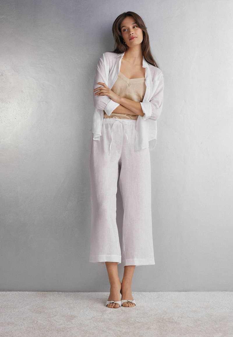 Linnen outfit met een lichtbeige tanktop en witte, losse, cropped broek. Vergezeld van witte sandalen met open tenen en accenten.