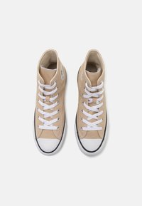 Beige plátěné vysoké tenisky s bílými tkaničkami a bílou špičkou. Mají černou gumovou podrážku a kulatou nášivku s logem na boku.