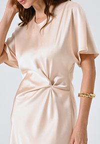 Donna che indossa un abito in satin beige chiaro con un dettaglio arricciato in vita e un grosso bracciale d'oro sul suo avambraccio destro.