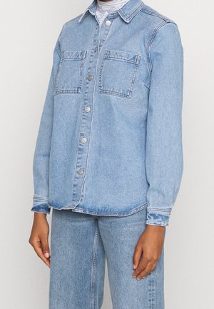 Osoba ubrana w jasnoniebieską kurtkę dżinsową z zapięciem na guziki, dwiema kieszeniami na piersi, zestawioną z pasującymi dżinsowymi spodniami oraz białym ażurowym topem pod spodem.