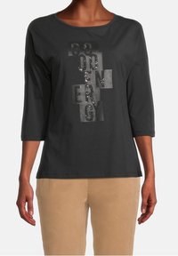 Schwarzes Langarmshirt mit rundem Ausschnitt; verfügt über einen glänzenden Textdesign mit Pailletten in verschiedenen Farbtönen; kombiniert mit beigen Hosen.