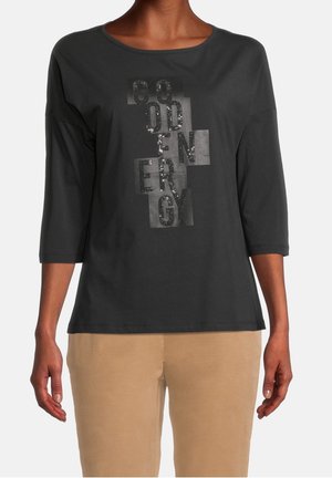 Schwarzes Langarmshirt mit rundem Ausschnitt; verfügt über einen glänzenden Textdesign mit Pailletten in verschiedenen Farbtönen; kombiniert mit beigen Hosen.