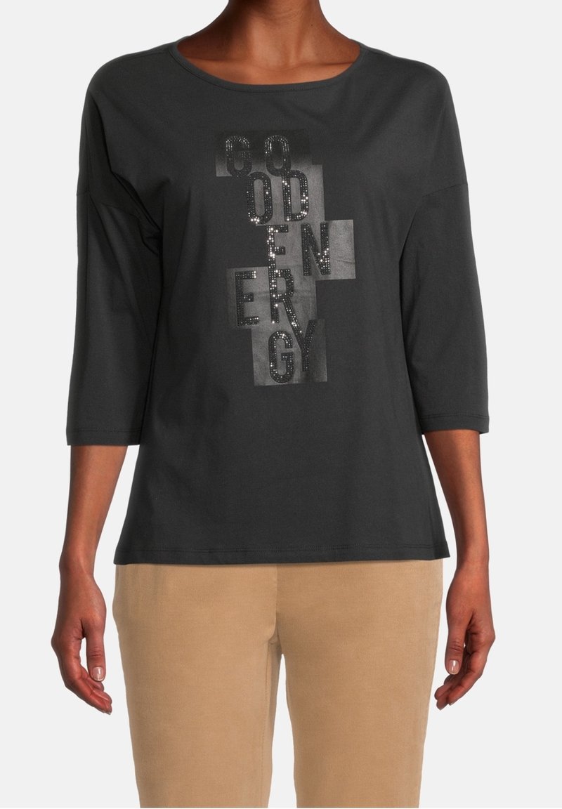 Schwarzes Langarmshirt mit rundem Ausschnitt; verfügt über einen glänzenden Textdesign mit Pailletten in verschiedenen Farbtönen; kombiniert mit beigen Hosen.