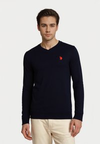 Joven con un suéter de cuello en V azul marino con un pequeño logo de caballo rojo y pantalones beige claros, de pie contra un fondo blanco puro.
