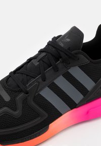 Svart idrottssko med en texturerad mesh-ovan, grått tre-randigt mönster, eleganta svarta detaljer och en livlig rosa-aprikos sula.