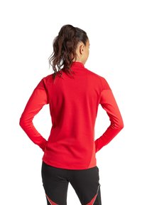 Rotes Langarm-Sportoberteil mit hohem Kragen und körperbetontem Design. Glatter Stoff mit genähten Nahtdetails. Kombiniert mit schwarzen Sporthosen.