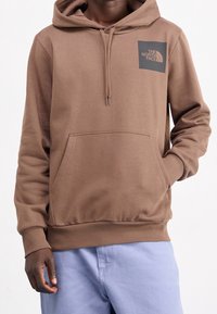 Osoba v hnědé mikině s kapucí s logem "The North Face" na hrudi a světle modrých kalhotách, jedna ruka v kapse, na jednobarevném pozadí.