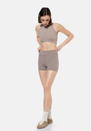 Kvinde justerer højtaljede taupe sportsshorts, iført matchende ærmeløst crop top, hvide sokker og beige sneakers på hvid baggrund.