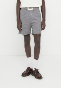 Σορτς με σχέδιο γκίνχαμ σε σκούρο μπλε και λευκό, με ελαστική μέση και ζώνη με λογότυπο. Συνδυάζονται με καφέ δερμάτινα παπούτσια και λευκές κάλτσες.