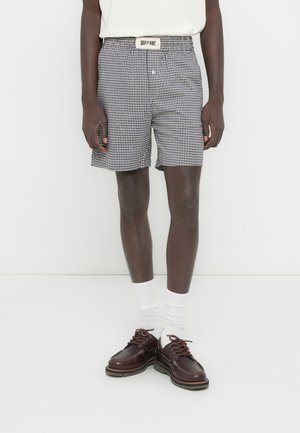 Σορτς με σχέδιο γκίνχαμ σε σκούρο μπλε και λευκό, με ελαστική μέση και ζώνη με λογότυπο. Συνδυάζονται με καφέ δερμάτινα παπούτσια και λευκές κάλτσες.