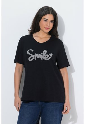 Kvinde med mørkt hår iført en sort T-shirt med grå tekstureret "Smile" tekst og blå jeans, stående mod en hvid væg.