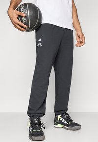 Pantalones deportivos gris oscuro con puños elásticos, decorados con detalles de logo en blanco. Combinados con zapatillas deportivas negras y sosteniendo un balón de baloncesto.