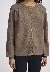 Ruskea neulecardigan, jossa on pyöreä kaula-aukko, pitkät hihat ja kuusi kultasävyistä nappia etupuolella. Pehmeä rakenne ja ribbireunat.